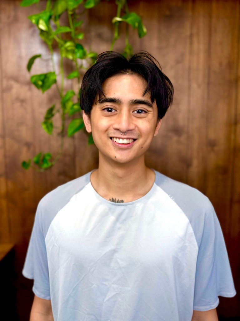 A young man with dark hair wearing a light blue shirt, smiling while standing in front of a wooden wall with green plants.